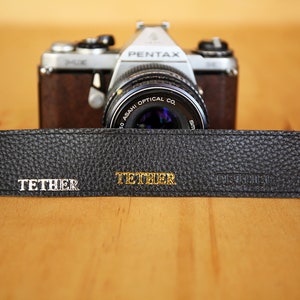 May include: A black leather camera strap with the word "TETHER" embossed in gold lettering. The strap is lying on a wooden surface in front of a silver camera with a black lens.
