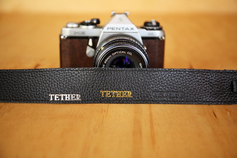 May include: A black leather camera strap with the word "TETHER" embossed in gold lettering. The strap is lying on a wooden surface in front of a silver camera with a black lens.