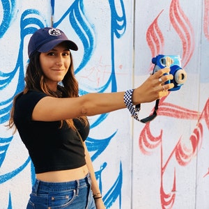 May include: A woman wearing a blue baseball cap with a white logo, a black crop top, and blue jeans, is holding a blue and white instant camera in front of a white wall with blue and red graffiti.