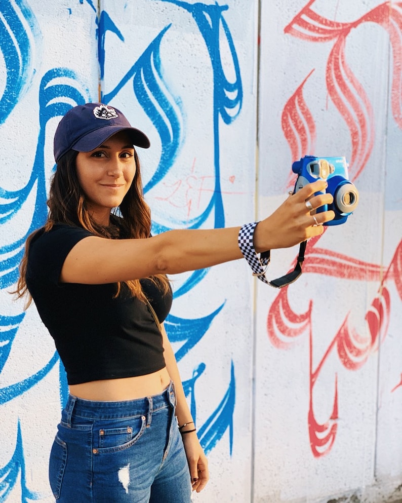 May include: A woman wearing a blue baseball cap with a white logo, a black crop top, and blue jeans, is holding a blue and white instant camera in front of a white wall with blue and red graffiti.