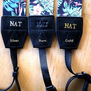 May include: Three camera straps with black leather ends and a floral print. The leather ends are personalized with the name "NAT" in silver, burn, and gold lettering.