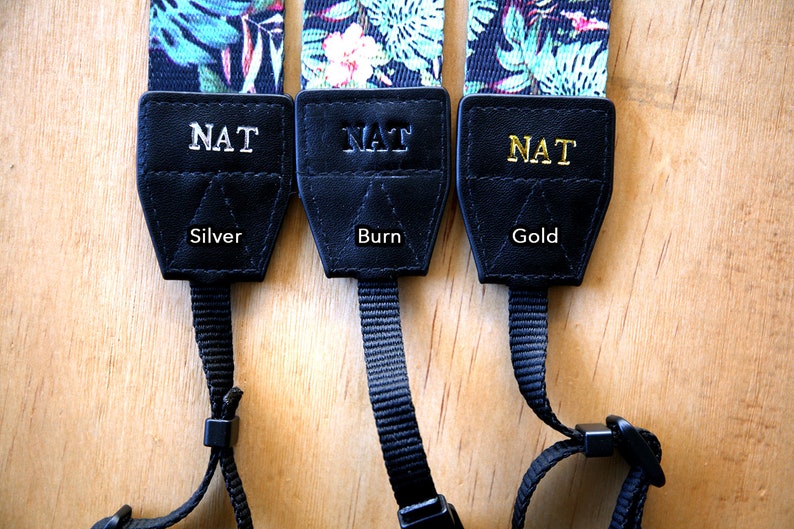 May include: Three camera straps with black leather ends and a floral print. The leather ends are personalized with the name "NAT" in silver, burn, and gold lettering.