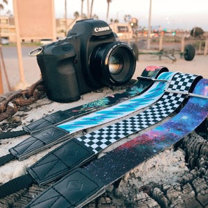 May include: Three camera straps with different patterns: a tropical floral print, a black and white checkered pattern, and a galaxy print. The straps are made of fabric and have black leather ends. A black Canon DSLR camera is in the background.