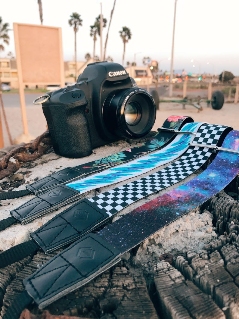 May include: Three camera straps with different patterns: a tropical floral print, a black and white checkered pattern, and a galaxy print. The straps are made of fabric and have black leather ends. A black Canon DSLR camera is in the background.