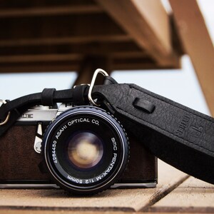 May include: A silver and brown 35mm film camera with a black leather strap. The camera lens is a 1:2.8 50mm SMC Pentax-M lens. The strap has the word "Artisan" embossed on it.