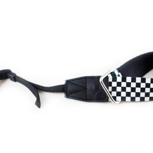 May include: Black and white checkered camera strap with a black leather adjustable section and a silver metal loop.