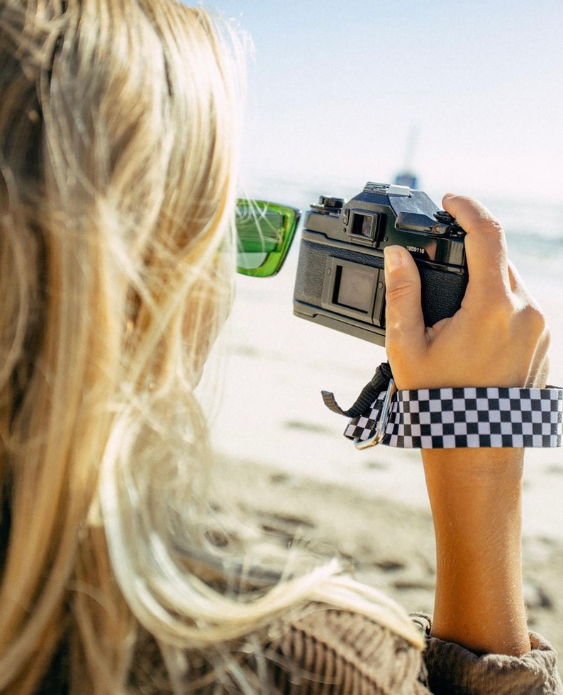May include: A person wearing green sunglasses holds a black camera with a checkered camera strap. The camera is pointed towards the horizon.