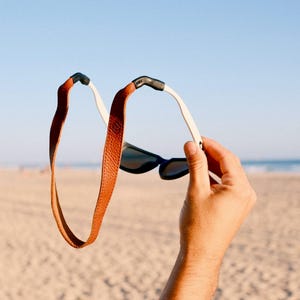 Peut inclure: Une paire de lunettes de soleil dotée d'une sangle en cuir texturé marron clair fixée aux branches blanches. La sangle forme une grande boucle s'étendant à l'arrière des montures aux verres sombres, avec des connecteurs en caoutchouc noir.