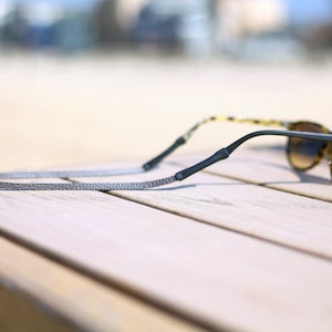 Peut inclure: Un cordon de lunettes en cuir gris avec des embouts en caoutchouc noir. Le cordon est enroul&eacute; autour d'une paire de lunettes de soleil en &eacute;caille de tortue.