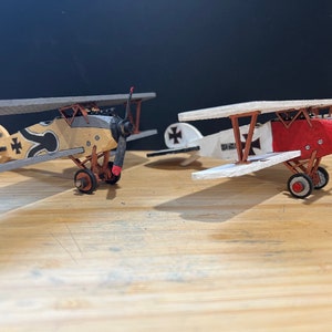 Puede incluir: Dos modelos de aviones, uno con un esquema de pintura rojo y blanco y el otro con un esquema de pintura marrón y gris. Ambos aviones tienen una cruz de hierro negra en las alas.