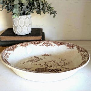 May include: An oval ceramic serving bowl featuring a cream-colored base with intricate brown transferware patterns. The rim displays geometric and floral motifs, while the center depicts a detailed scene of flowering branches and a fan.