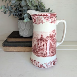 May include: A cream-colored ceramic pitcher featuring a deep red transferware pattern. The design displays an intricate architectural scene with arched structures and surrounding foliage, complemented by floral borders at the top and base rims.