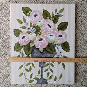 May include: An original painting of a floral arrangement in a gray vase. The flowers are pink and white, with green leaves and blue accents. The background is striped with beige and white. The painting is approximately 16 inches wide.