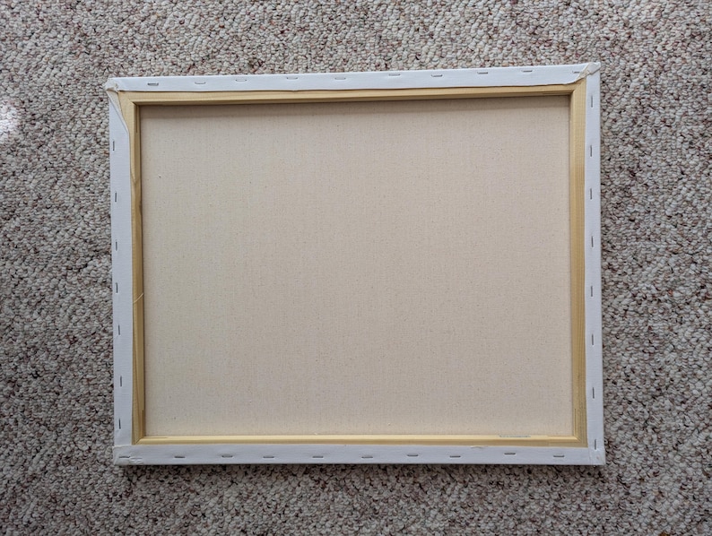 May include: Back of a rectangular canvas stretched over a wooden frame. The canvas is a light beige color, and the frame is a natural wood tone. The canvas is stapled to the wooden frame. The edges of the canvas are white.