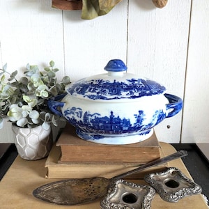 May include: A white ceramic tureen with a blue transferware pattern depicting a landscape scene, featuring two side handles and a matching lid with a knob. The tureen is accompanied by a tarnished silver-plated cake server and two square candle holders.