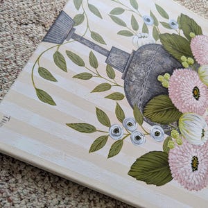 May include: A floral painting featuring a gray watering can surrounded by pink and white flowers, green leaves, and small blue flowers. The background has vertical beige and white stripes. The artwork is on a canvas.