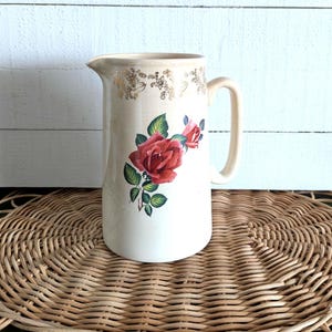 May include: Cream-colored ceramic pitcher with a red rose floral design. The pitcher has a handle and a spout. Gold floral accents adorn the rim. The pitcher is displayed on a woven wicker placemat.