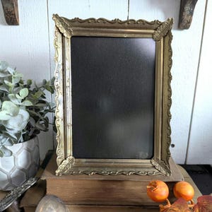 May include: A rectangular brass picture frame with an ornate, scalloped edge and decorative floral corner accents. The frame features a smooth, dark, solid-colored insert panel, providing a stark contrast to the metallic, weathered gold-toned finish of the metal border.