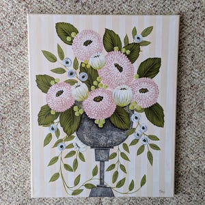 May include: An original painting of a floral arrangement in a gray urn. The flowers are pink and white with green leaves, set against a striped background. The artwork is signed in the lower right corner.