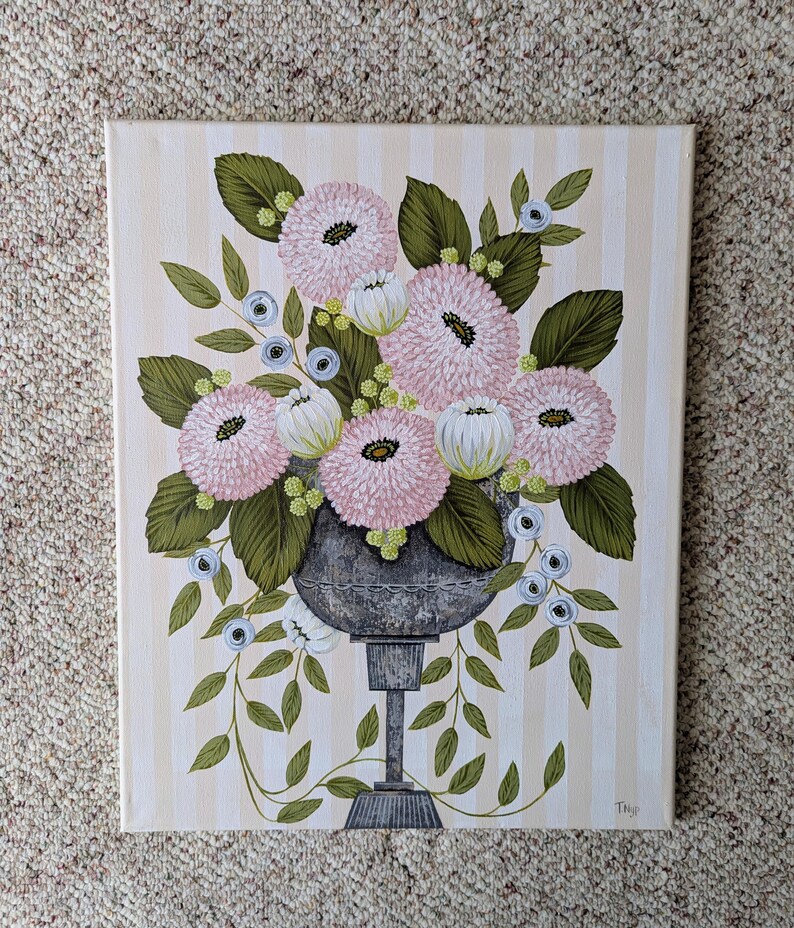 May include: An original painting of a floral arrangement in a gray urn. The flowers are pink and white with green leaves, set against a striped background. The artwork is signed in the lower right corner.