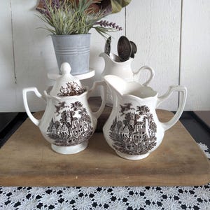 May include: A cream-colored ceramic sugar bowl with a lid and a matching creamer pitcher, both featuring a detailed brown transferware landscape print of buildings and trees. The pieces have scalloped rims and curved handles, showcasing a classic design.