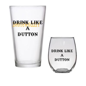 May include: Two clear glass drinking glasses with the text "Drink Like A Dutton" printed on them. The text is in black with a yellow outline. The larger glass is a pint glass and the smaller glass is a wine glass.