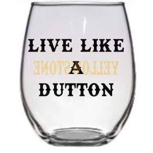 May include: A clear glass wine glass with the text "LIVE LIKE A YELLOWSTONE DUTTON" printed on it. The text is in black and gold.