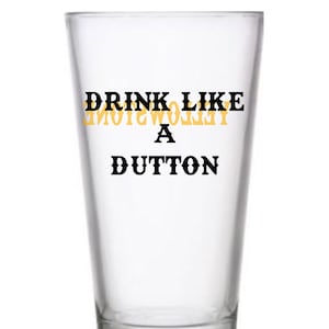 May include: A clear glass drinking glass with the text "Drink Like A Yellowstone Dutton" printed on it in black and yellow.