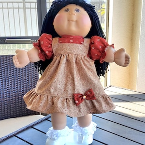 May include: A vintage doll with black hair and a round face, wearing a red and brown dress with a bow. The doll has white shoes with lace trim and is standing on a dark surface. The doll's arms are outstretched.
