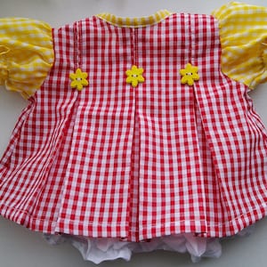 May include: A red and white gingham baby dress with yellow floral accents. The dress features yellow ruffled sleeves and three yellow flower-shaped buttons down the back. The dress has a white underlayer.