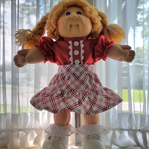 Puede incluir: Una muñeca con cabello de lana rubia peinado en coletas, que viste una blusa roja de manga corta con lunares blancos y tres botones blancos, combinada con una falda a cuadros blancos, rojos y negros, calcetines con encaje y zapatos blancos.