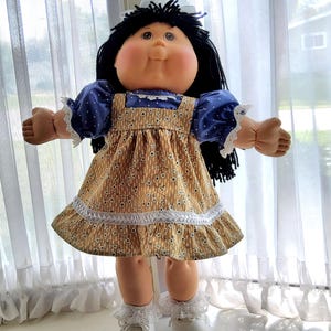 Könnte beinhalten: Eine Vintage-Puppe mit schwarzen Haaren und rundem Gesicht. Die Puppe trägt ein gold-blau gemustertes Kleid mit weißer Spitzenborte und blauen Ärmeln. Die Arme der Puppe sind ausgestreckt.