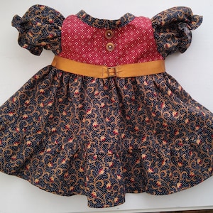 Puede incluir: Vestido de muñeca con corpiño burdeos y falda azul marino. El corpiño tiene un pequeño patrón de puntos blancos repetidos y dos botones marrones. La falda presenta un diseño floral con detalles dorados y rojos. Un cinturón color canela con hebilla ciñe la cintura.
