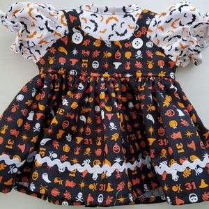 May include: A black pinafore dress featuring an orange and white Halloween print with ghosts, pumpkins, skulls, and the number 31, layered over a white shirt with a black bat pattern. The dress has two white buttons and white rickrack trim.