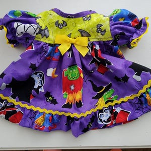 May include: A purple dress featuring a Halloween-themed print with vampires, Frankenstein's monster, witches, bats, and spiderwebs. The dress has a lime green yoke with a bat pattern, yellow ruffled trim, and a yellow bow at the center chest.