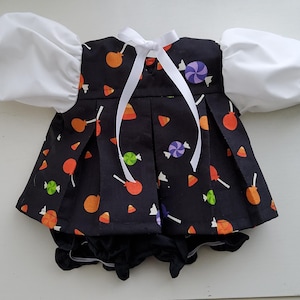May include: A black baby outfit with a candy print, featuring orange, green, and purple lollipops and candy corn. The outfit has white puff sleeves and a white bow. The skirt is pleated and the shorts are black.
