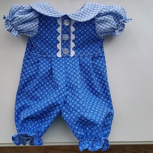 Op de afbeelding: Een blauwe romper met een wit cirkelpatroon, korte pofmouwen met een lichtblauw en wit gingham ruitpatroon, een bijpassende gingham kraag en een centrale verticale witte geschulpte bies met drie ronde witte knopen.