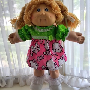 May include: A doll with blonde yarn hair styled in pigtails, featuring blue eyes and dimpled cheeks. The doll wears a green short-sleeved top with white polka dots and a pink skirt patterned with Hello Kitty graphics, white lace trim, and white shoes.
