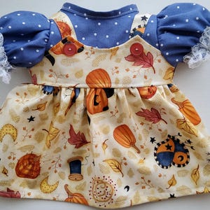 May include: A doll dress featuring a cream-colored skirt with a print of orange pumpkins, autumn leaves, stars, moons, and thread spools. The bodice has two red buttons, and the blue puff sleeves are decorated with white polka dots and white lace trim.