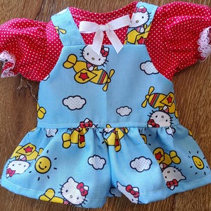 May include: A doll dress featuring a blue romper with a Hello Kitty airplane print, red polka dot sleeves, and white lace trim. A white bow adorns the neckline. The dress is designed for a doll.