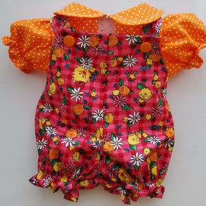 May include: A doll outfit consisting of a short-sleeved orange shirt with white polka dots and a pink gingham romper featuring a floral pattern with yellow and white flowers. The romper has two orange buttons at the top and ruffled leg openings.