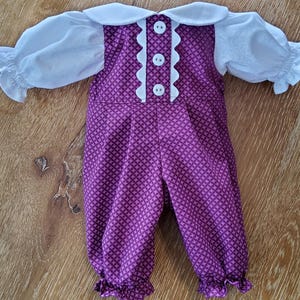 May include: A doll's romper with a white collar and sleeves. The romper is a deep purple with a repeating geometric pattern. The collar and sleeves are white. Three white buttons adorn the front. The romper has ruffled cuffs at the ankles.
