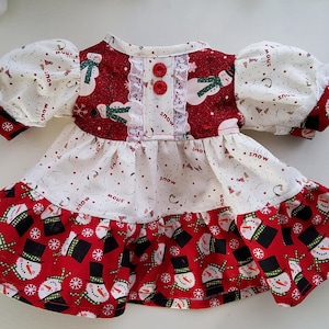 Può includere: Un vestito da neonato festivo a tema pupazzo di neve. L'abito presenta una combinazione di colori rosso e bianco con motivi di pupazzi di neve e fiocchi di neve. Ha maniche corte e a sbuffo ed è adornato con bottoni rossi e finiture in pizzo.