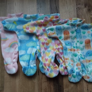 May include: Four fleece baby sleepsuits in pastel shades. Each suit has a unique pattern: pink with rainbows, blue with clouds, pink with hearts, and white with sea creatures. They have a zip-up front and footed design.