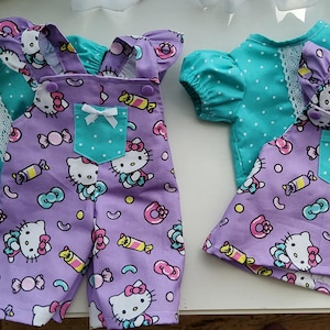 May include: Two doll outfits: a purple overall set and a dress, both with a Hello Kitty print. The overalls have a teal pocket and white bow. The dress has a teal pocket and a white lace trim. Both outfits have teal polka dot tops.