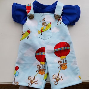 Op de afbeelding: Een lichtblauwe tuinbroek met een patroon van aapjes in heteluchtballonnen en vliegtuigen, gedragen over een effen koningsblauw shirt met korte mouwen. De tuinbroek heeft twee metalen knopen op de schouderbanden en een enkele borstzak.