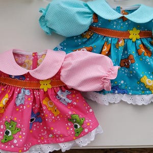 Könnte beinhalten: Zwei Puppenkleider, eines rosa und eines blau, mit Care Bear-Mustern. Jedes Kleid hat einen Bubikragen, Puffärmel und eine weiße Spitzenborte. Das rosa Kleid hat einen rosa Kragen und Ärmel, während das blaue Kleid einen blauen Kragen und Ärmel hat.