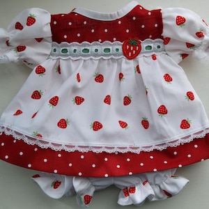 May include: A doll dress and matching bloomers set featuring a white fabric with a red strawberry print and a red fabric with white polka dots. The dress has puff sleeves with white lace trim, a white lace waistband, and a strawberry applique.