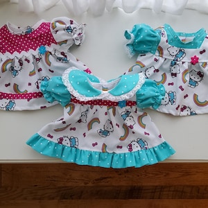 May include: Three doll dresses featuring a white fabric base with a repeating pattern of Hello Kitty, rainbows, and small bows. The dresses include puff sleeves, colorful polka-dot accents in magenta or teal, and small decorative flower-shaped buttons.