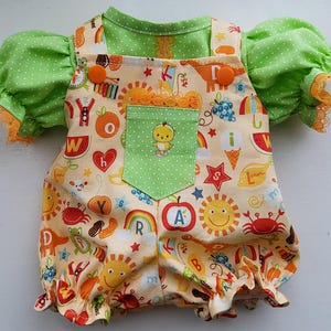 May include: A light green polka dot shirt with orange lace trim on the sleeves paired with cream-colored overalls featuring a colorful print of letters, fruits, suns, and animals. The overalls have a green pocket with a small yellow chick graphic.